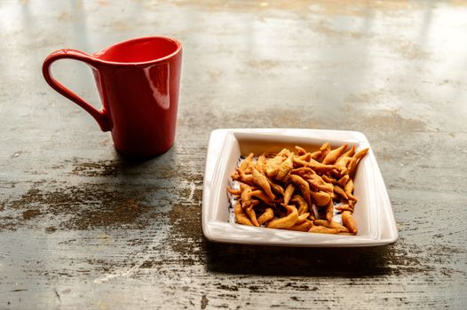 Close up Red Color Coffee mug and white plate full of Namkeen Bhujia snack cookies on old rustic floor. Food and drink background. Morning sunlight coming from window.