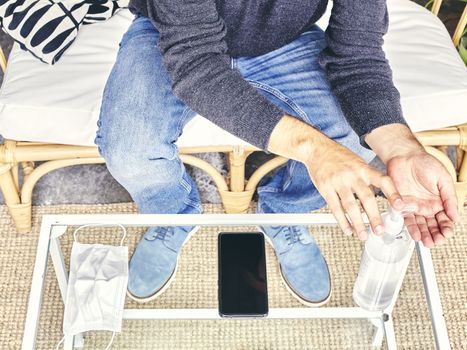 Man in jeans sitting on a sofa wiping his hands with hydroalcohol gel and with his mask and phone resting on the glass table