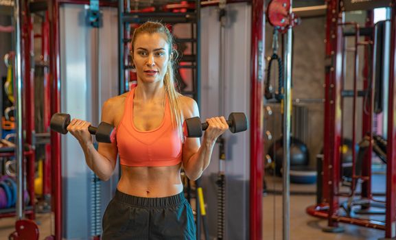 young woman exercising with dumbbells to strengthen biceps and shoulders in gym.
