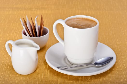 Mug of coffee with saucer, teaspoon, milk jug and sugar sachets on a golden table cloth 