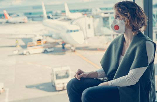 Woman with a mask on her mouth protects against the virus. She looks sad through the window at the airport on planes. Aerial connections canceled due to a coronavirus.