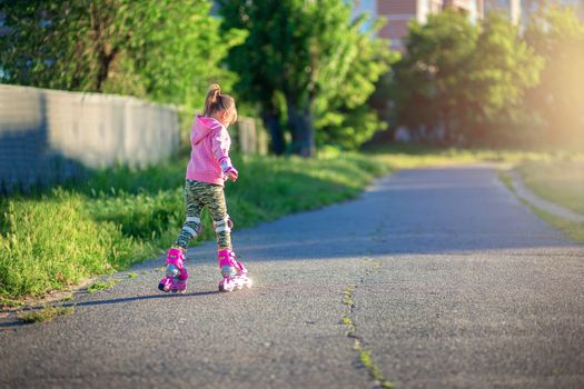 A little girl rides on pink roller skates on asphalt in the summer. Photo without a face from the back