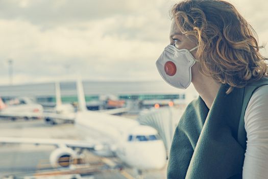 Woman with a mask on her mouth protects against the virus. She looks sad through the window at the airport on planes. Aerial connections canceled due to a coronavirus.