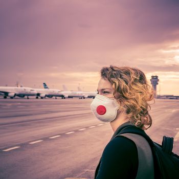 Woman with a mask on her mouth protects against the virus. She looks sad through the window at the airport on planes. Aerial connections canceled due to a coronavirus.