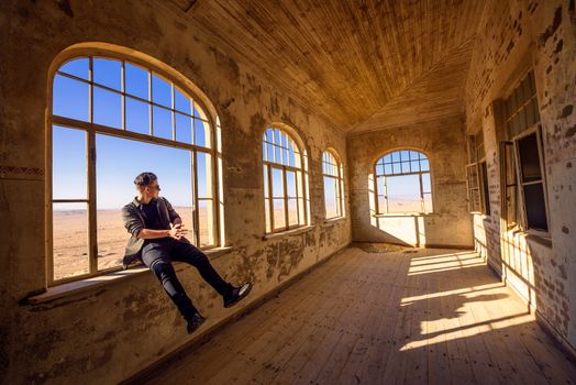 Tourist sits in a widow of a ruined house located in the mining town Kolmanskop. Kolmanskop is a ghost town in the Namib desert near Luderitz in Namibia, Southern Africa.