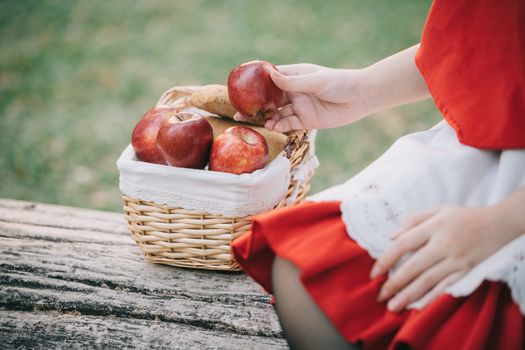 Portrait young woman with Little Red Riding Hood costume with apple and bread on basket sitting in green tree park background