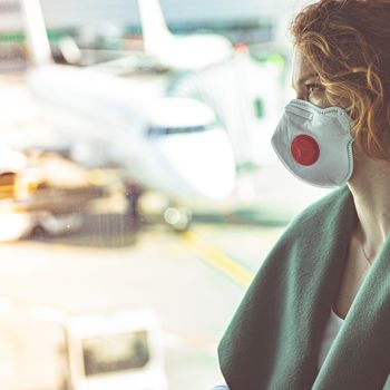 Woman with a mask on her mouth protects against the virus. She looks sad through the window at the airport on planes. Aerial connections canceled due to a coronavirus.