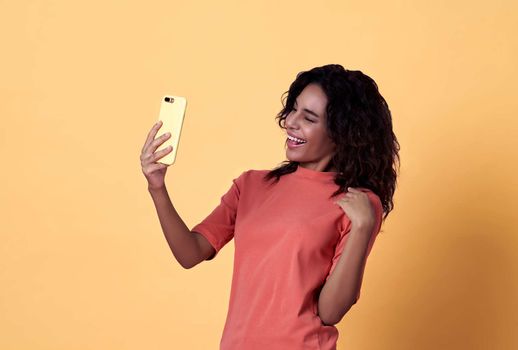 Portrait of a happy young african woman selfie celebrating with mobile phone isolated over yellow background.