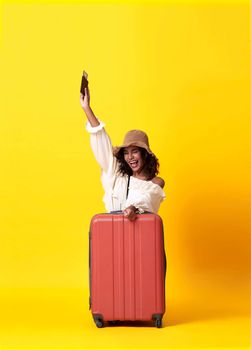 Cheerful young african woman dressed in summer clothes holding passport with suitcase over yellow background.