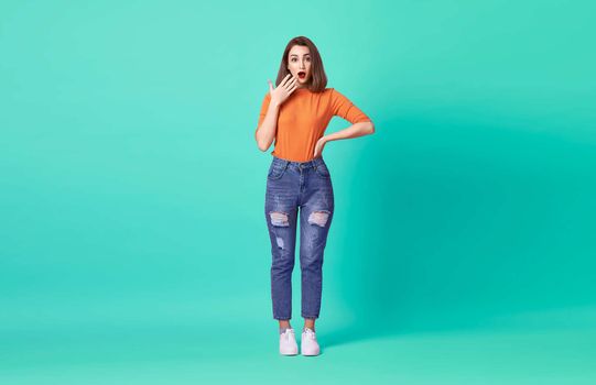 Shocked excited beautiful woman with mouth open wearing casual orange t-shirt over blue background.