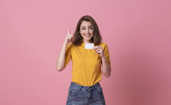 Young smiling beautiful woman in yellow shirt showing credit card in hand over pink background.