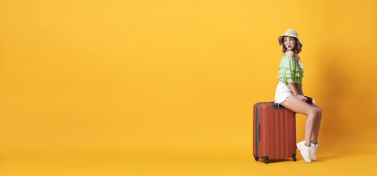 Cheerful young woman dressed in summer clothes holding passport with suitcase over yellow banner background with copy space.