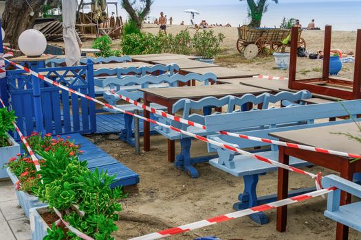 Closed outdoor bar restaurants with stacked chairs, tables & no access tape with bathers on sand by the sea.