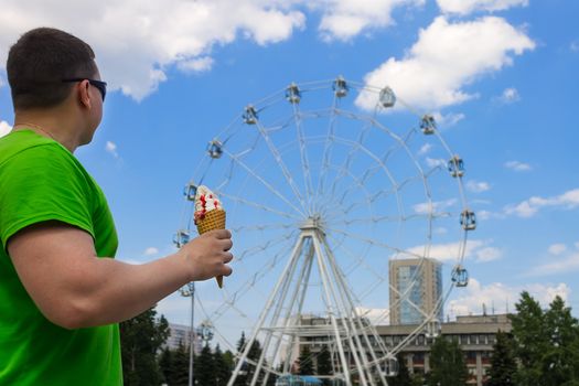 The guy in green clothes holds ice cream in his hand in the park leisure and entertainment on the background of the Ferris wheel