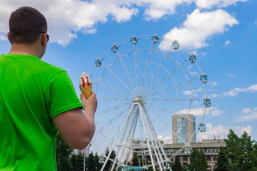 The guy in green clothes holds ice cream in his hand in the park leisure and entertainment on the background of the Ferris wheel