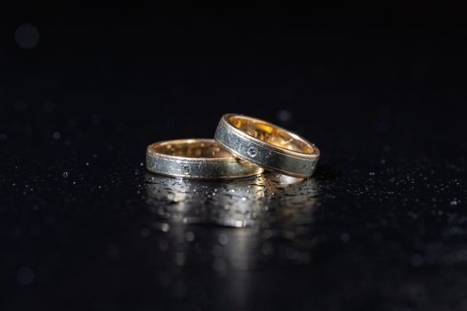 Wedding rings lying on dark surface shining with light close up macro. Water splashes