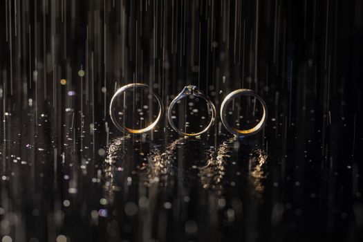 Wedding and engagement rings lying on dark surface shining with light close up macro. Water splashes. Rain