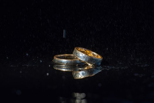 Wedding rings lying on dark surface shining with light close up macro. Water splashes