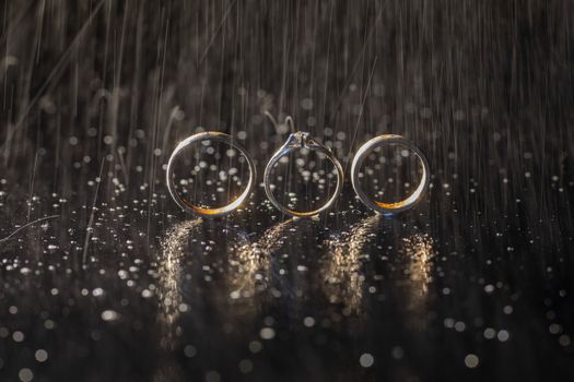 Wedding and engagement rings lying on dark surface shining with light close up macro. Water splashes. Rain