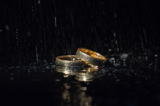 Wedding rings lying on dark surface shining with light close up macro. Water splashes