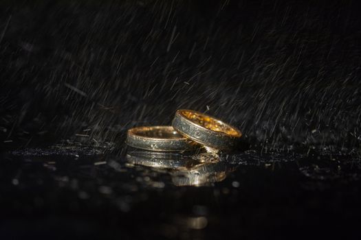 Wedding rings lying on dark surface shining with light close up macro. Water splashes