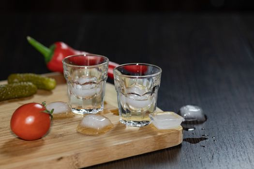 Two shot glasses of vodka on the cutting board with cherry tomatoes, marinated cucumbers, pepper and ice cubes. Black background