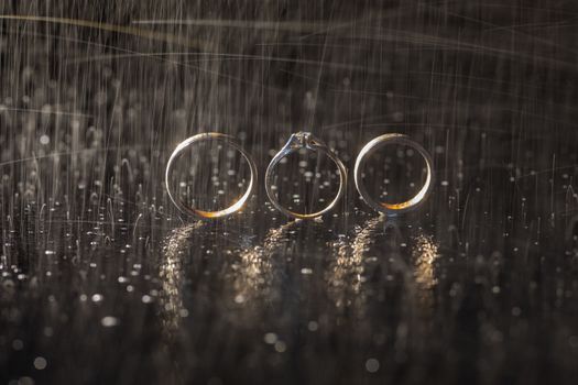 Wedding and engagement rings lying on dark surface shining with light close up macro. Water splashes. Rain