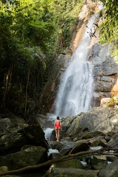 Beautiful young slim blonde woman with curly hair, wearing in red swimsuit is enjoy in lagoon of huge tropical waterfall in jungle. Travel concept. Sexy woman wearing red swimsuit in the jungle