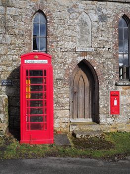 Traditional red British public telephone box containing a defibrillator unit standing outside a chapel converted into a telegraph office with a postbox in the wall, Dartmoor National Park, Devon, UK
