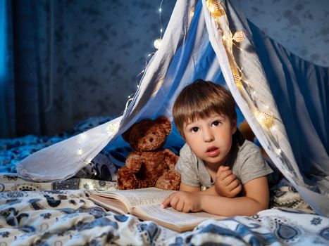 Little boy reads book. Toddler plays in tent made of linen sheet on bed. Cozy evening with favorite book.