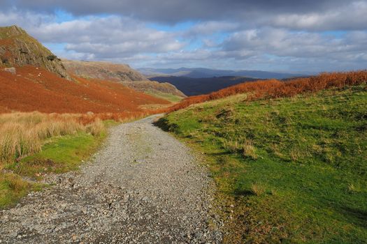 Path over the mountains through heather and bracken in warm Autumn orange colour under a blue and white cloudy sky, Lake District, UK
