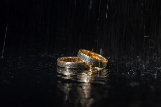 Wedding rings lying on dark surface shining with light close up macro. Water splashes