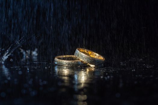 Wedding rings lying on dark surface shining with light close up macro. Water splashes