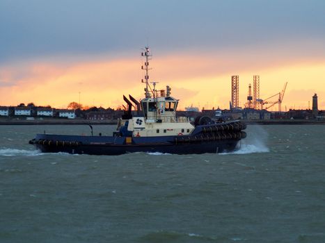 Harbor tug from the Port of Felixstowe makes its way back up the river against the background of an orange sunset overlooking the cranes and seafront of Harwich, Suffolk, UK