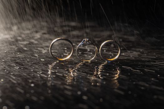 Wedding and engagement rings lying on dark surface shining with light close up macro. Water splashes. Rain