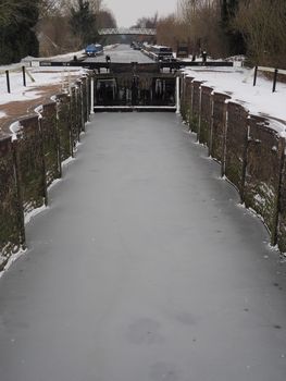 Winter snow and frozen ice in the bottom of the historic Aldermaston Lock with canal boats and footbridge in the distance, Kennet and Avon Canal, Berkshire, UK