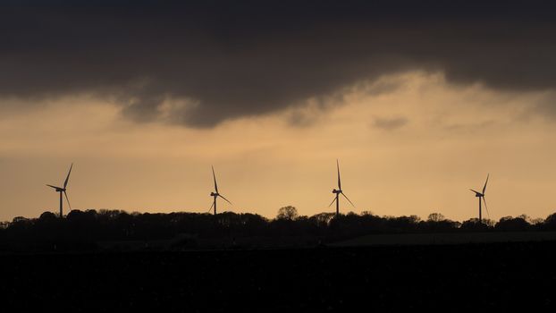 Wind turbines on a hillside silhouetted against a stormy and overcast evening sky at sunset