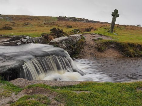 Windy Post granite cross standing behind a waterfall cascading over rocks on the Grimsbridge and Sortridge leat under a grey sky, Dartmoor National Park, Devon, UK