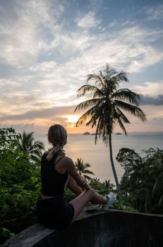 Silhouette of woman sitting alone on the hill with a coconut palm trees and enjoys sea on a horizon in a sunset. Vacation and travel concept on a island. Beautiful sunsets on a Samui island