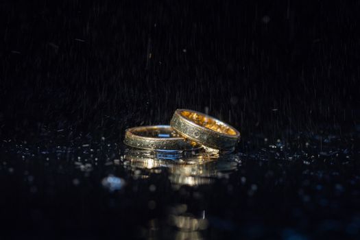 Wedding rings lying on dark surface shining with light close up macro. Water splashes