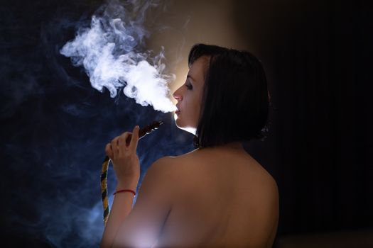 Beautiful, young woman smoking hookah. Attractive girl smoking flavored tobacco. Blow out smoke on black background. Shooting from the back of the model