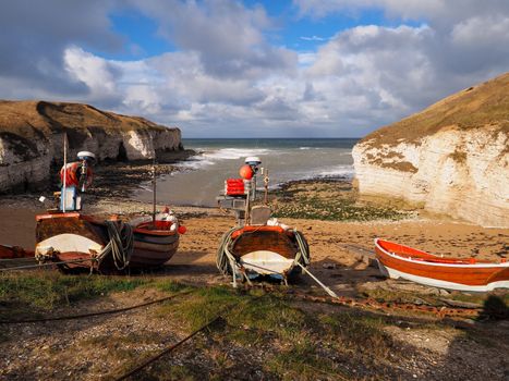 Colorful wooden and red and white fishing boats tied up on the beach in a cove waiting for the tide to come in with white cliffs in the background and a blue sky with clouds overhead.
