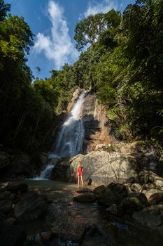 Beautiful young slim blonde woman with curly hair, wearing in red swimsuit is enjoy in lagoon of huge tropical waterfall in jungle. Travel concept. Sexy woman wearing red swimsuit in the jungle