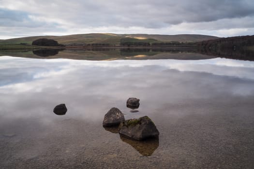 The serene clear water of Malham Tarn with the reflection of the hills in the water and the late afternoon sun highlighting the hills behind, Yorkshire Dales, UK