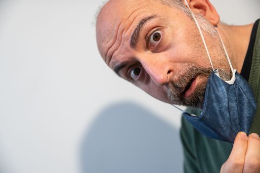 Portrait of an extremely perplexed middle-aged Caucasian man. The head is tilted and the gaze fixed on the lens with eyes wide with amazement. With one hand he lowered the mask for the coronavirus