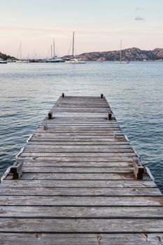 Wooden pier and sailboats mooring in background at evening calm sea of marvellous Porto Rafael, Costa Smeralda, Sardinia, Italy. Symbol for relaxation, wealth, leisure activity.