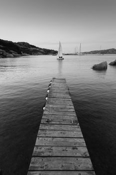 Wooden pier and sailboats sailing in evening calm sea of marvellous Porto Rafael, Costa Smeralda, Sardinia, Italy. Symbol for relaxation, wealth, leisure activity. Black and white image.