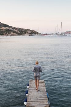 Beautiful woman in luxury summer dress standing on wooden pier enjoying peaceful seascape at dusk. Female traveler stands on a wooden pier in Porto Rafael, Costa Smeralda, Sardinia, Italy.