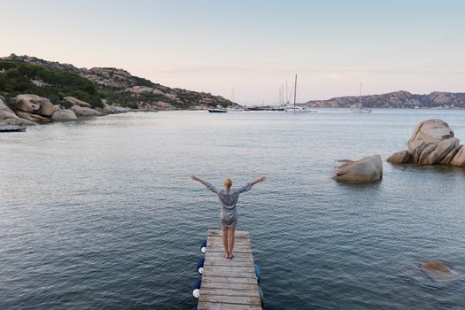 Beautiful woman standing on wooden pier arms rised enjoying peaceful seascape at dusk. Female traveler stands on a wooden pier in Porto Rafael, Costa Smeralda, Sardinia, Italy.