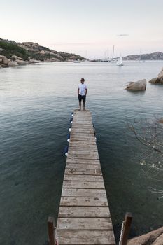 Man standing on wooden pier enjoying peaceful seascape at dusk. Male tourist stands on a wooden pier in Porto Rafael, Costa Smeralda, Sardinia, Italy.